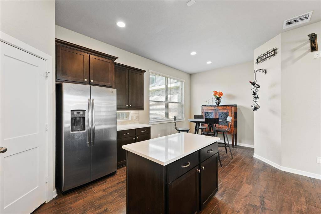 a modern kitchen with dark wood cabinets, stainless steel appliances, and a large white countertop island with pendant lighting. The open layout leads to a dining area and a staircase with a dark wood railing. The space is bright and inviting, with ample natural light.