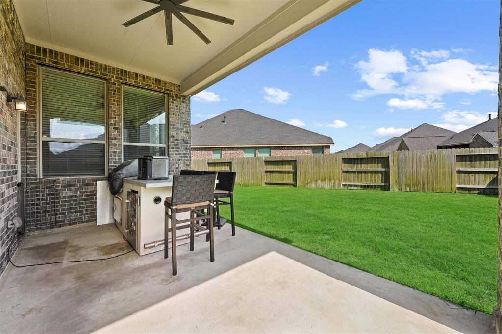 a covered patio with a ceiling fan and an outdoor kitchen setup, overlooking a well-maintained grassy backyard enclosed by a wooden fence. Ideal for outdoor entertaining and relaxation.