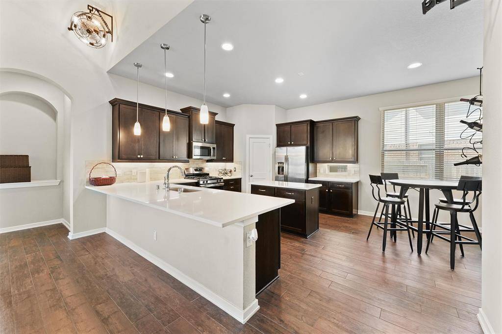 This modern kitchen features dark wood cabinets, stainless steel appliances, and a large island with pendant lighting. The open layout includes a dining area with a high table, all set against rich, wood-look flooring. Ideal for both cooking and entertaining.