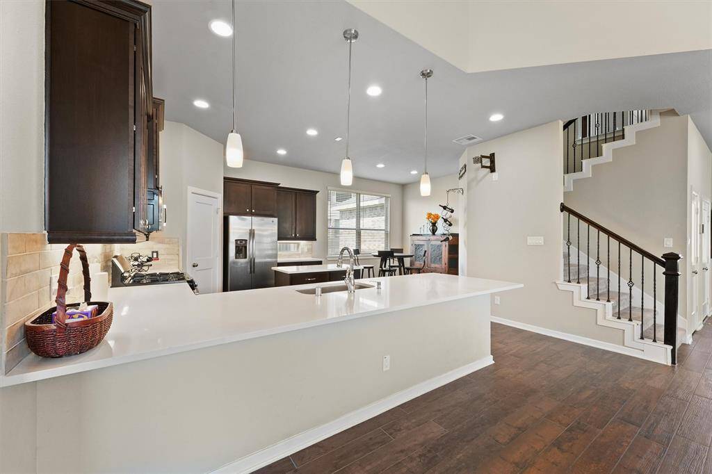 a modern kitchen with dark wood cabinets, stainless steel appliances, and a large white countertop island with pendant lighting. The open layout leads to a dining area and a staircase with a dark wood railing. The space is bright and inviting, with ample natural light.