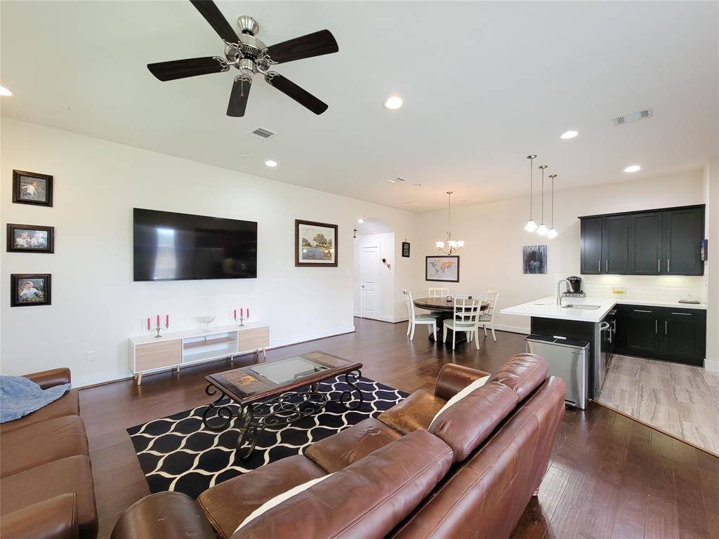 Modern living room with adjoining kitchen with a dining area; neutral tones with wood flooring and ceiling fan.