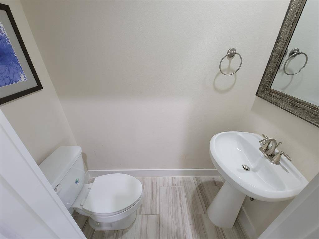 A clean, modern bathroom with a toilet, pedestal sink, mirror, towel rings, and a framed picture on the wall.