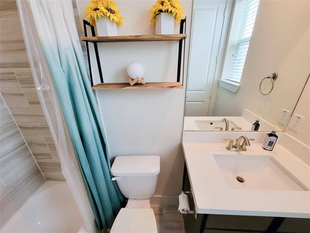 A clean bathroom with a blue shower curtain, white toilet, sink, decorative shelves with yellow flowers, and a wood-textured tile floor.