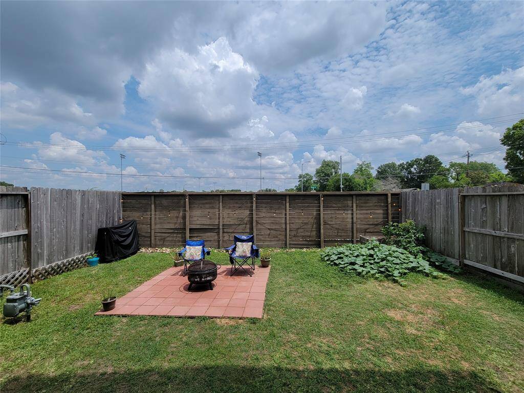 A backyard with a lawn, two chairs, a barbecue grill on a tile surface, plants, a wooden fence, and a partly cloudy sky above.