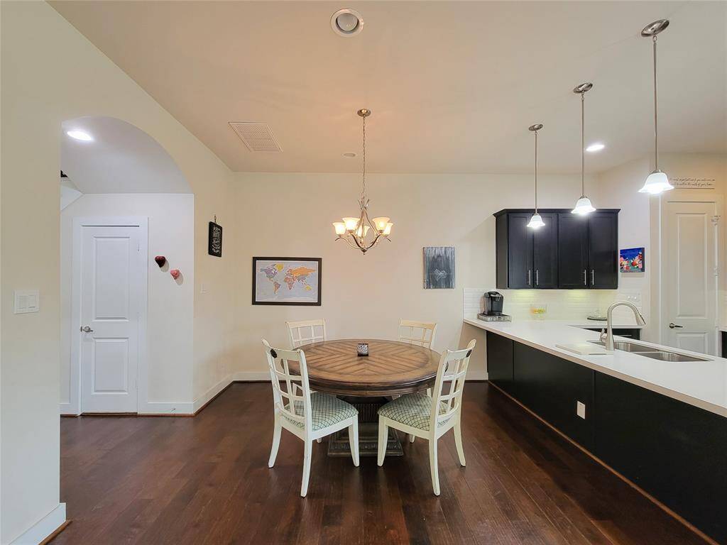 A modern kitchen with wooden floors, white walls, a dining table, pendant lights, and black cabinets.