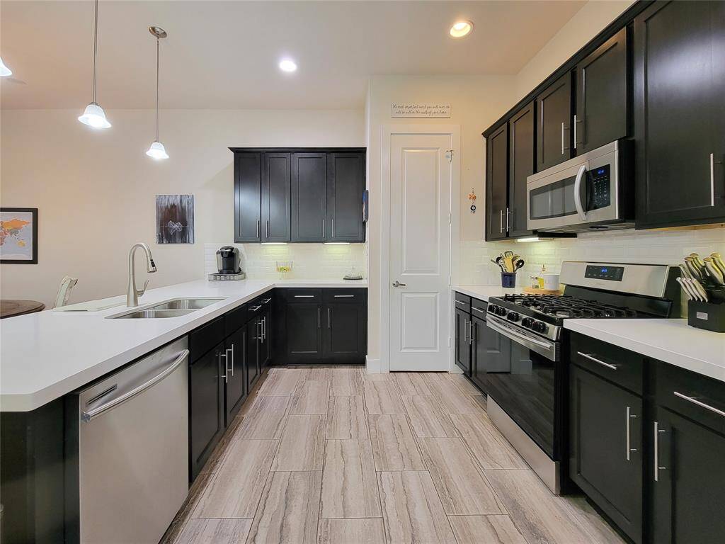 A modern kitchen with dark cabinets, stainless steel appliances, white countertops, and pendant lighting over a wood-look floor.