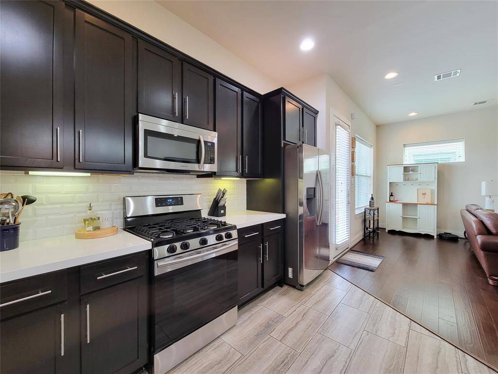 Modern kitchen with dark cabinets, stainless steel appliances, white backsplash, and hardwood floors, leading to a bright living area with furniture.