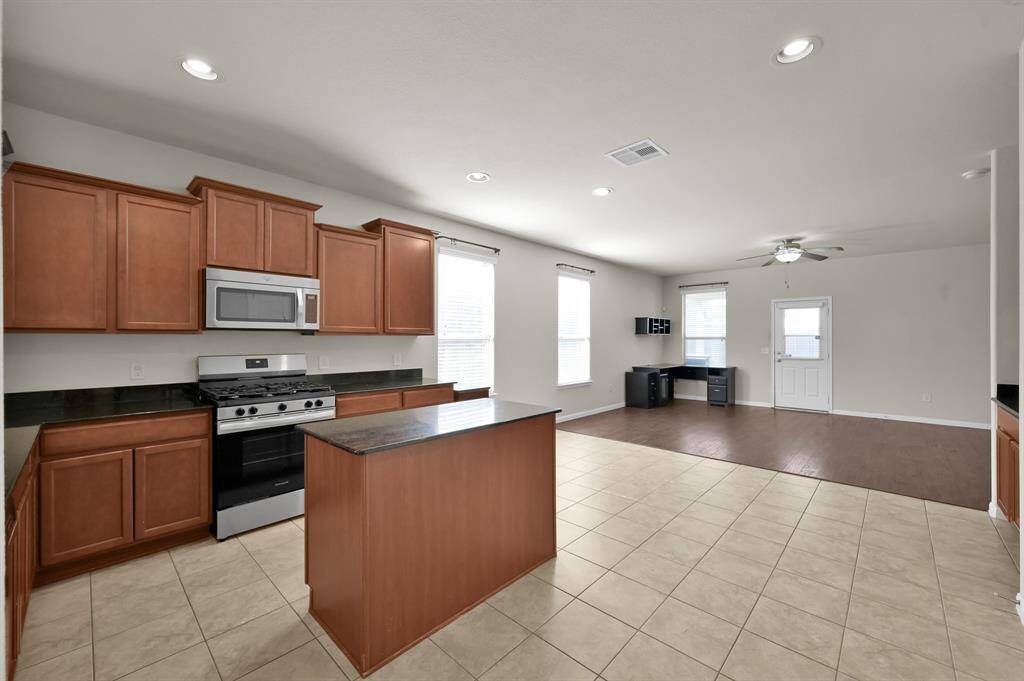 Spacious, modern kitchen with tile flooring, ample natural light, and an open layout. It features stainless steel appliances, wooden cabinetry, and a central island, leading into a well-lit living area.