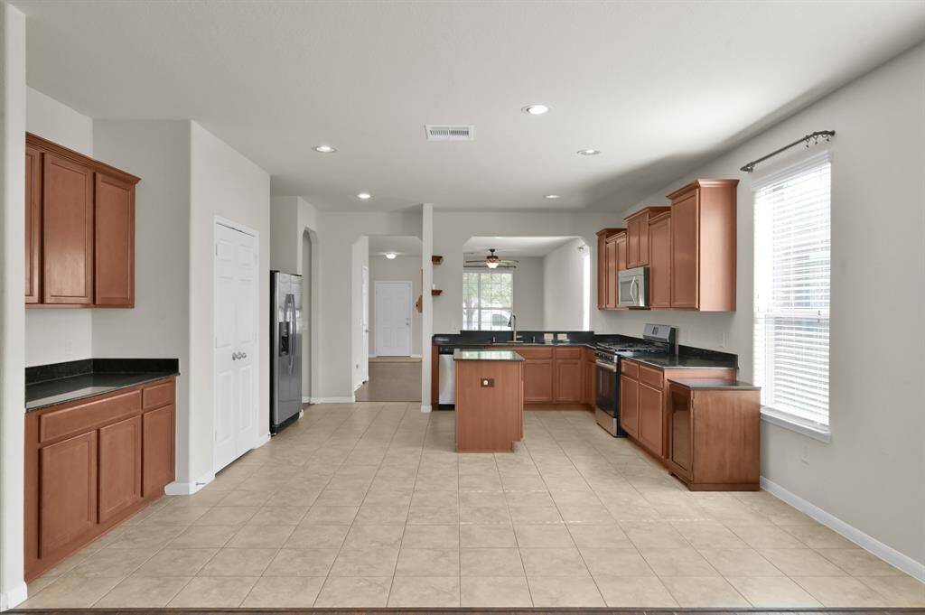 Kitchen with modern stainless steel appliances, granite countertops, and wooden cabinetry. It features tile flooring, an island, and an open layout that connects to a bright living area with hardwood floors.