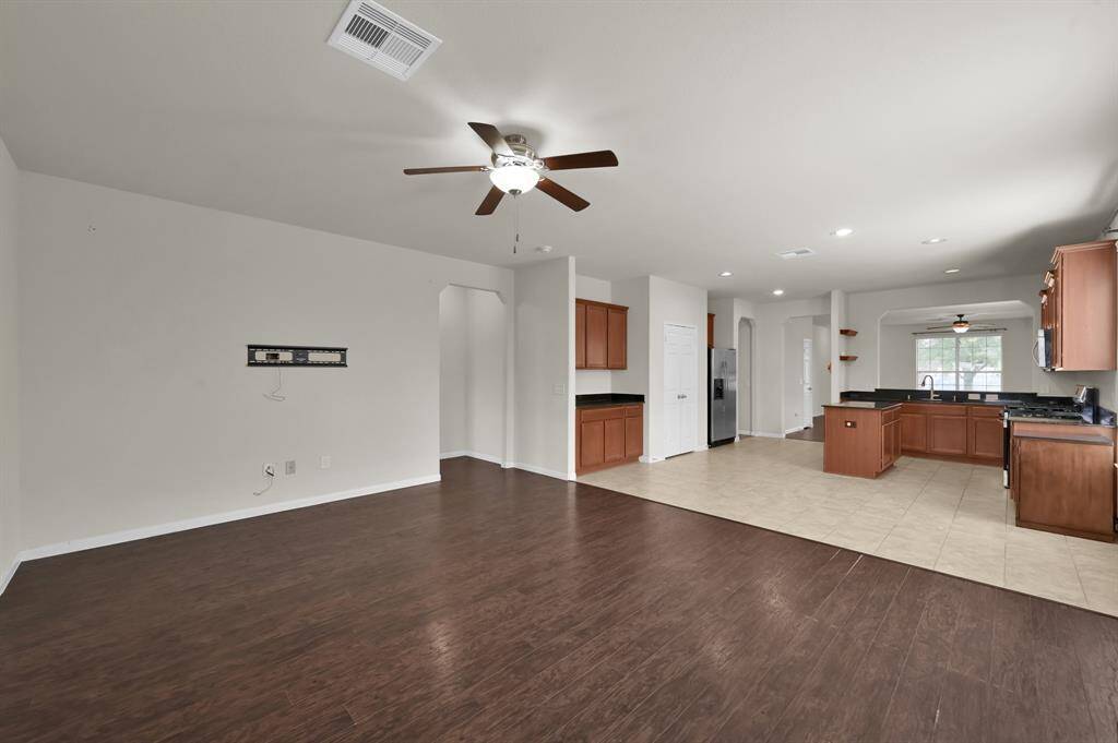 This spacious, open-plan living area features hardwood floors, a ceiling fan, and natural light from large windows. It seamlessly connects to a modern kitchen with stainless steel appliances and tile flooring, offering a perfect blend of comfort and functionality.