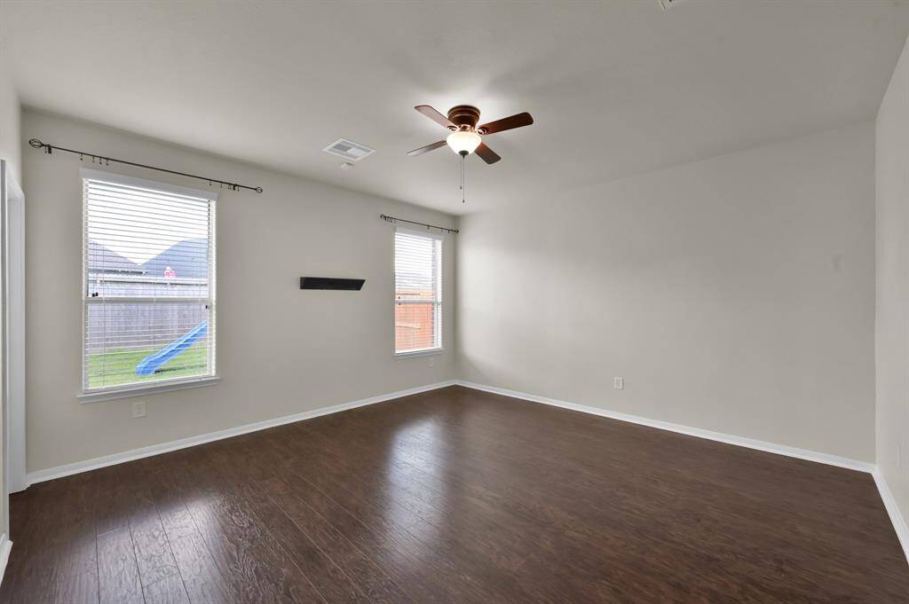 Dark wood flooring and neutral walls. It features a ceiling fan, two doors, and a wall mount for a TV. Large windows provide ample natural light, and there's an entrance to an adjacent bathroom.