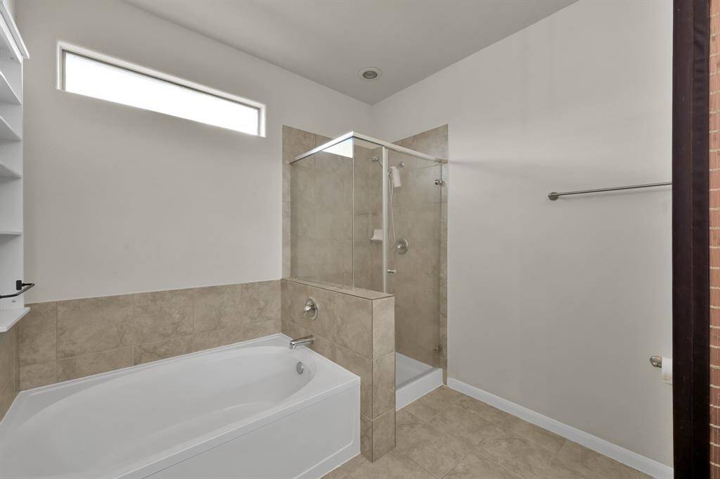 This bathroom features a modern design with a spacious bathtub and a separate glass-enclosed shower. It has neutral tiles, a horizontal window for natural light, and a towel bar.