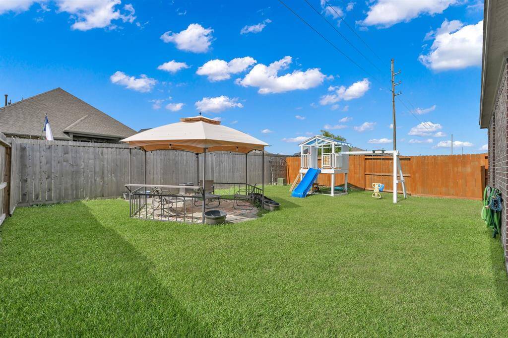 This backyard features a covered patio area with seating, a well-maintained lawn, a swing set, and a brick exterior. It is fully fenced, providing privacy and a safe space for outdoor activities.