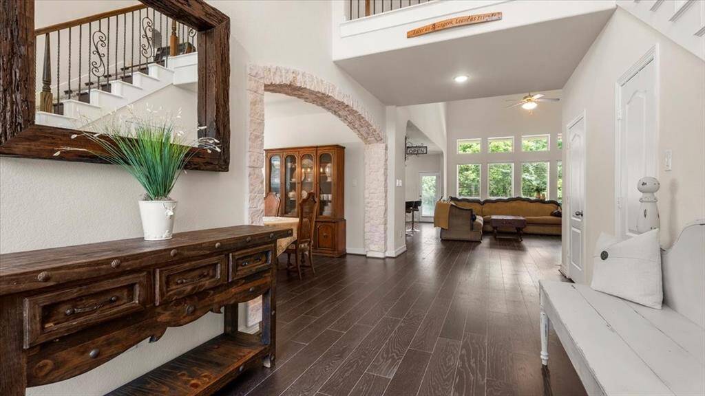 This inviting home offers an open-concept layout with a warm, spacious living area featuring dark wood flooring, soaring ceilings, and abundant natural light streaming through large windows. A stone archway leading to the formal dining room and an elegant staircase add timeless charm and architectural interest.