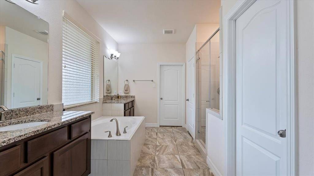 The primary bathroom offers a spacious layout with his-and-hers sinks set in granite countertops, a sleek soaking tub, and a separate glass-enclosed shower. Neutral tile flooring and abundant natural light contribute to the bright, spa-like ambiance.
