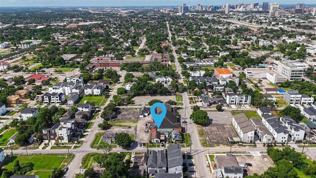 This aerial photo showcases a residential neighborhood with a mix of modern and traditional homes, lush greenery, and tree-lined streets. A blue marker highlights the specific property in a vibrant urban setting, with cityscape views in the distance, indicating convenient proximity to downtown amenities.