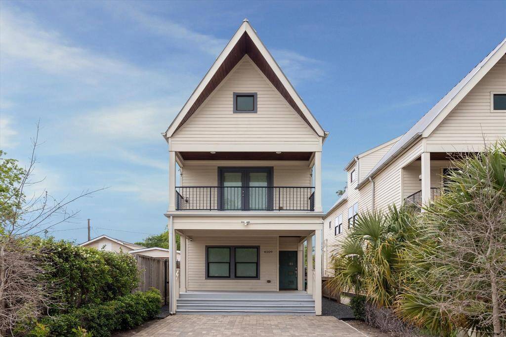 This is a modern, two-story home with a steeply pitched roof and a covered balcony. It features a neutral exterior, small front porch, and a paved driveway. The property is flanked by greenery for added privacy.