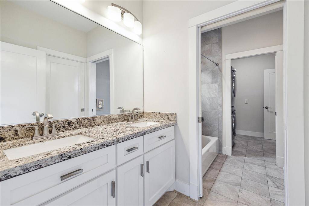 This bathroom features a modern double vanity with granite countertops, sleek faucets, and ample cabinetry. It includes a shower-tub combo with tiled walls and a view into an adjacent room with stacked laundry appliances. The neutral tones create a clean, inviting space.