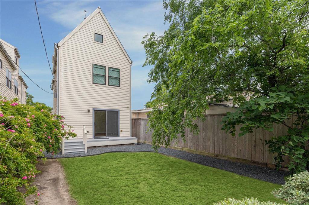 This photo shows a charming, narrow two-story house with a simple, modern exterior. The backyard features a small, well-maintained lawn, surrounded by a wooden fence and mature trees, providing privacy and shade. A sliding glass door opens to a small deck, perfect for outdoor relaxation.