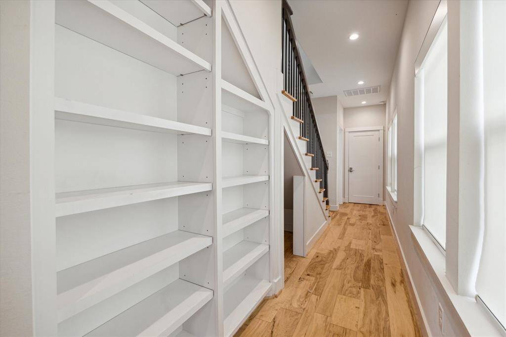 This hallway features built-in white shelves, a modern staircase with black railings, and light wood flooring. It is brightened by natural light from large windows, creating an inviting and functional space.
