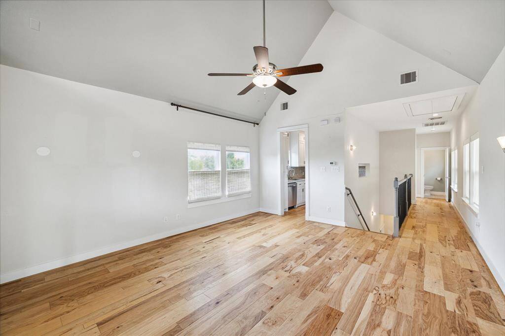 This inviting room features high vaulted ceilings, beautiful hardwood floors, and abundant natural light from large windows. A ceiling fan enhances comfort, and there's a cozy nook leading to a kitchen area. The space is open and airy, ideal for comfortable living.
