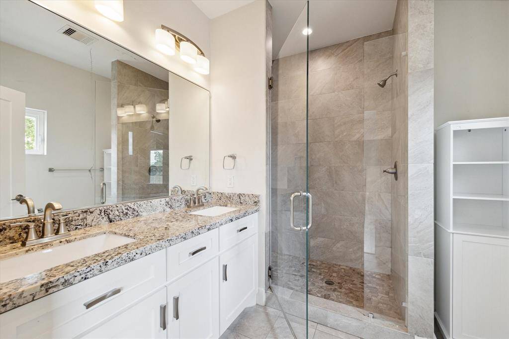 This bathroom features a modern design with a spacious glass-enclosed shower, dual sinks with granite countertops, and sleek white cabinetry, offering ample storage and a clean aesthetic.