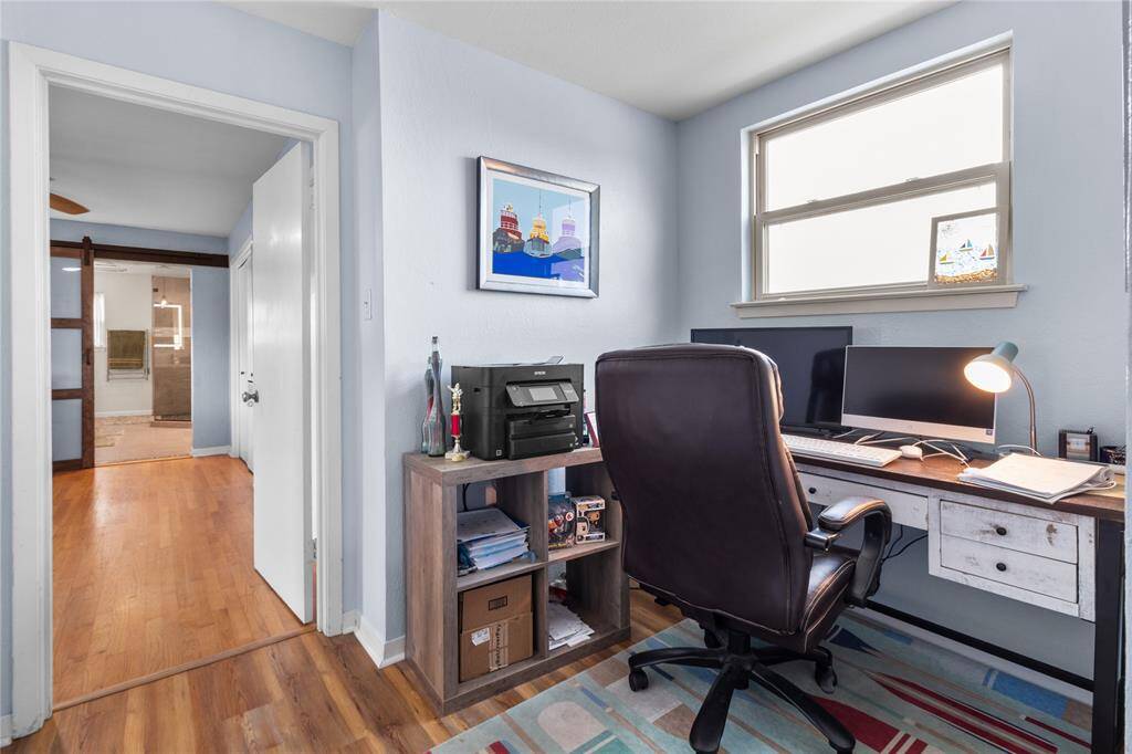 This photo shows a cozy home office with light blue walls, a large window, and hardwood floors. The room connects to a hallway leading to another area of the house.