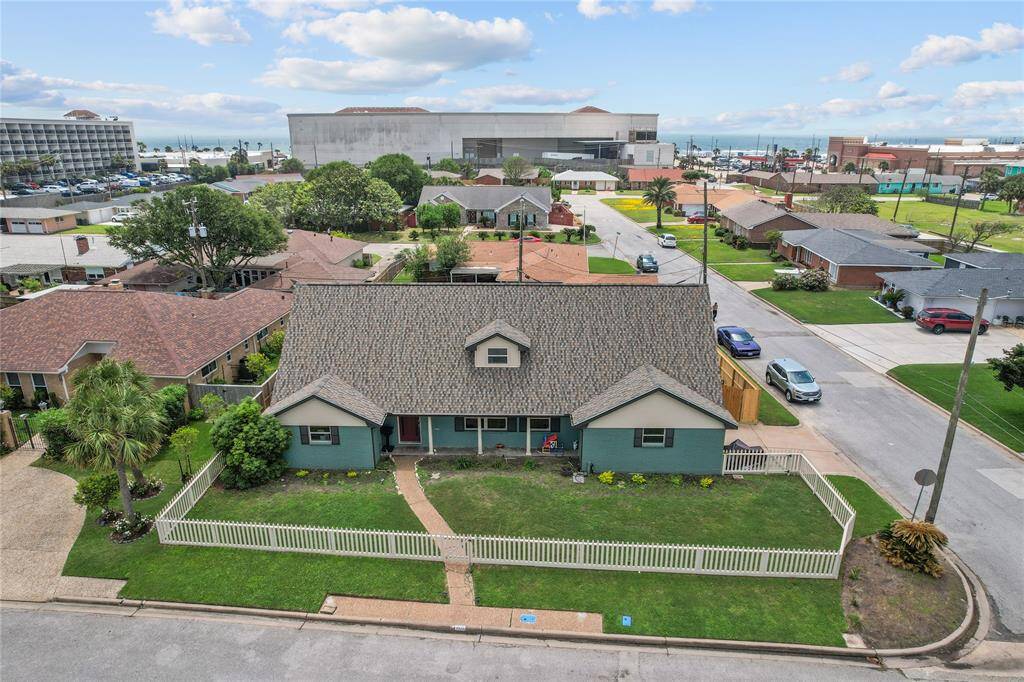 This charming home features a well-maintained exterior with a green facade and a neatly manicured lawn, enclosed by an off white picket fence. It is situated on a corner lot in a residential neighborhood, offering convenient proximity to nearby amenities and a view of the ocean in the background.